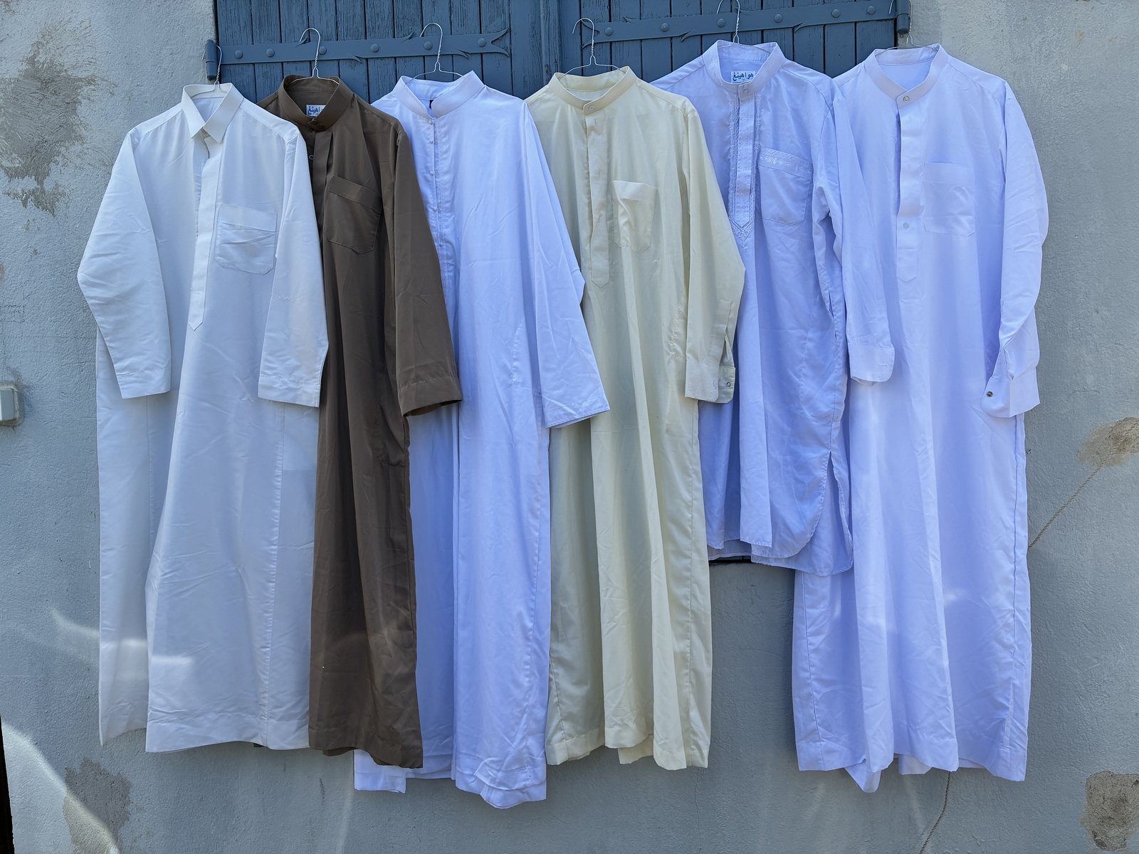 Six qamis et jellabas sur cintres dans des coloris blanc, brun, bleu, écru, lilas et lavande