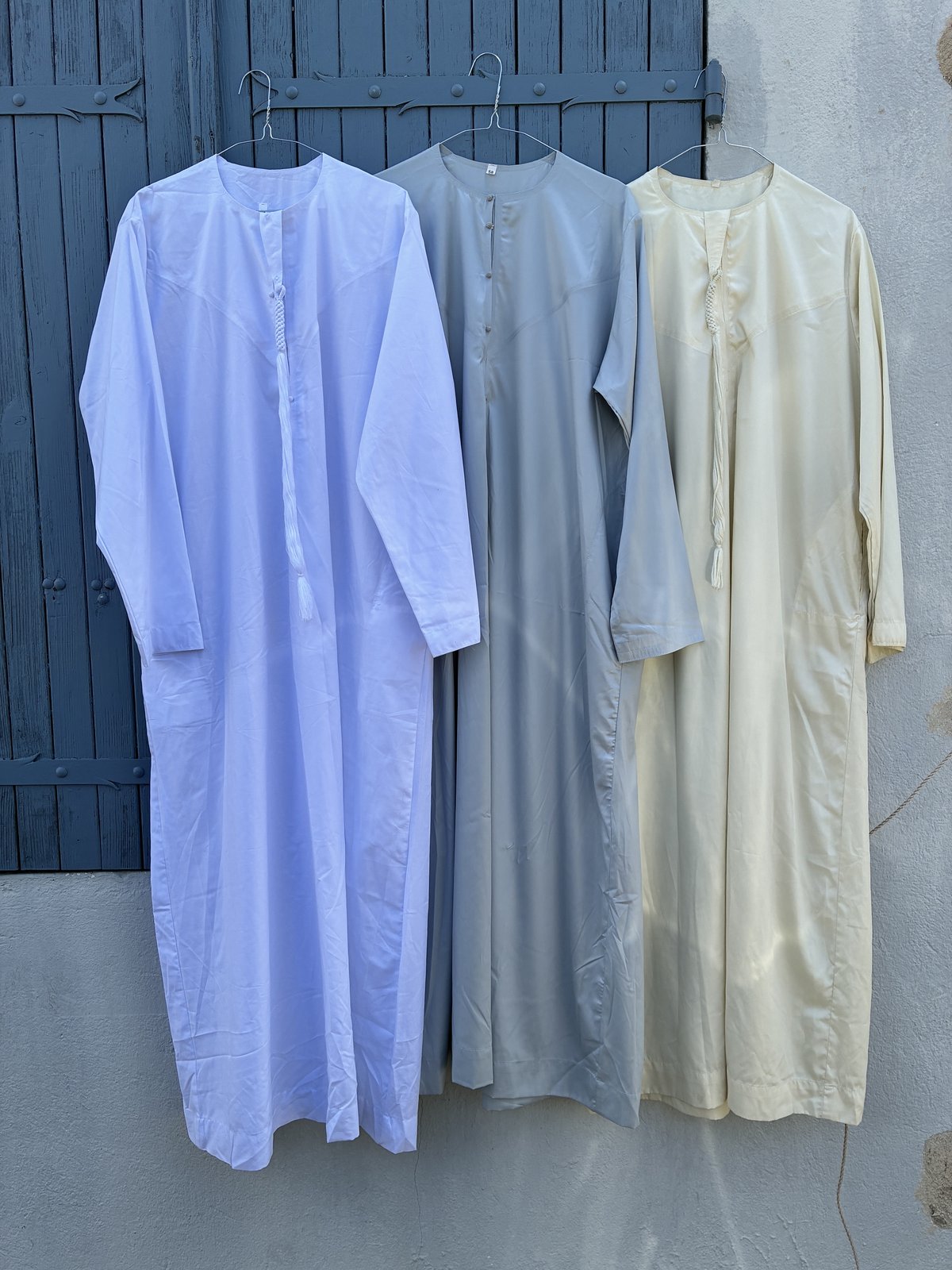 Trois longues robes traditionnelles à col fermé en crème, gris et blanc, vues à plat