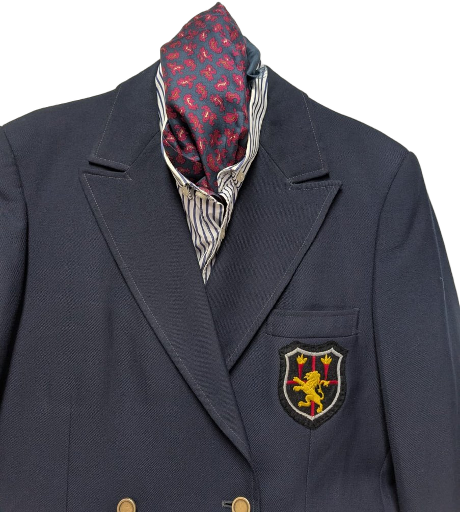 Détail du blazer college : écusson héraldique brodé (lion d'or rampant, flèches), foulard ascot bordeaux à motifs paisley sur chemise à fines rayures bleues