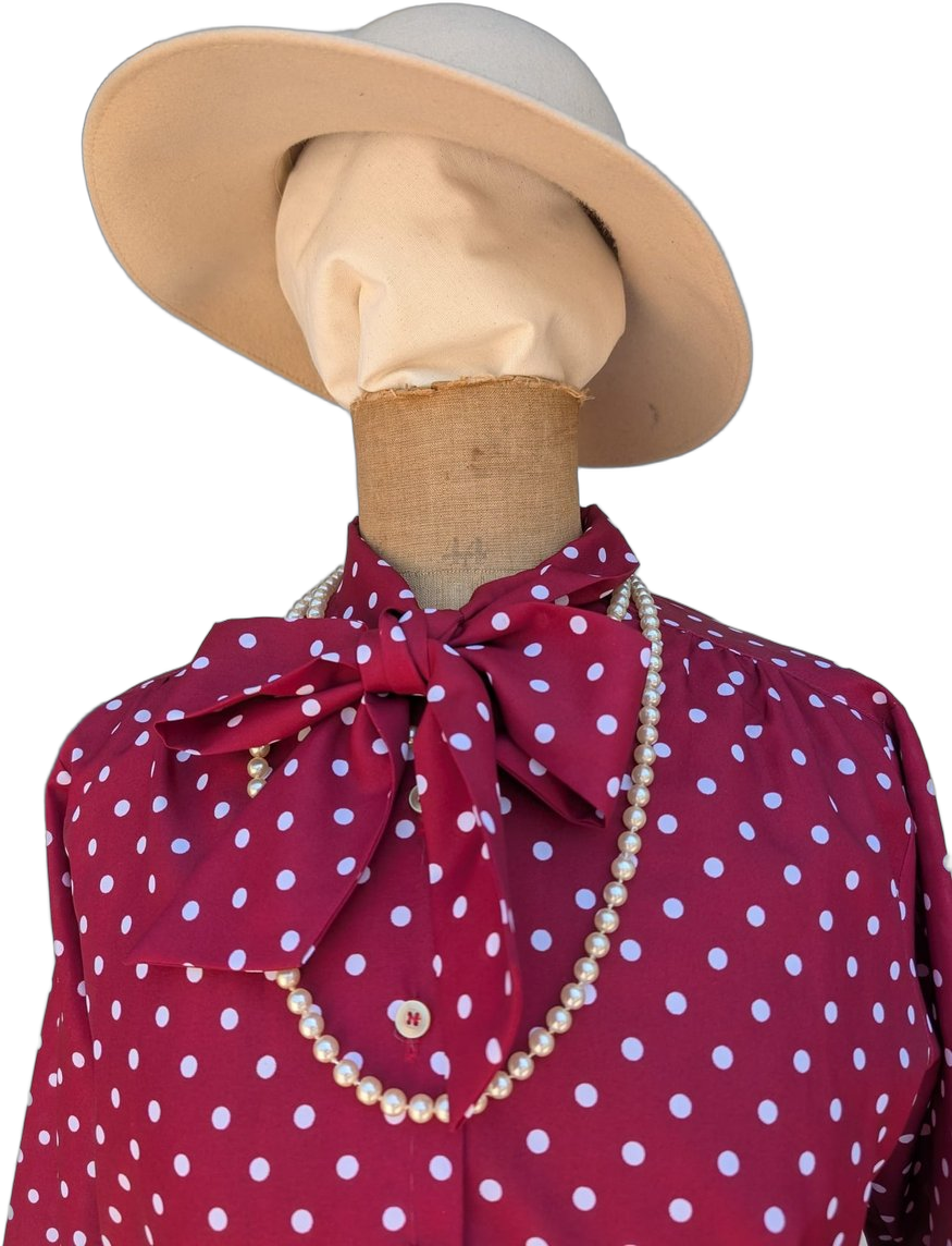 Détail buste : capeline beige large bord, lavallière à pois bordeaux nouée, collier de perles double rang sur chemisier à pois